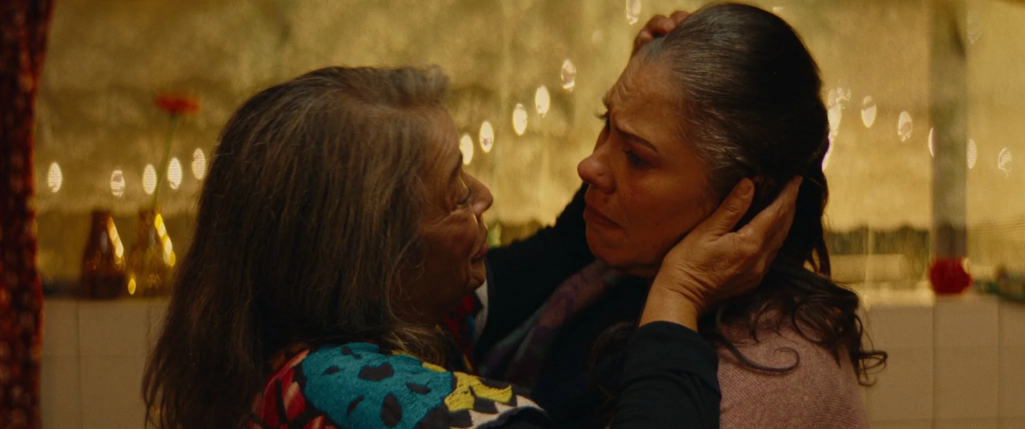 Two women in warm candlelight, one cradling the other’s face as they reconcile.