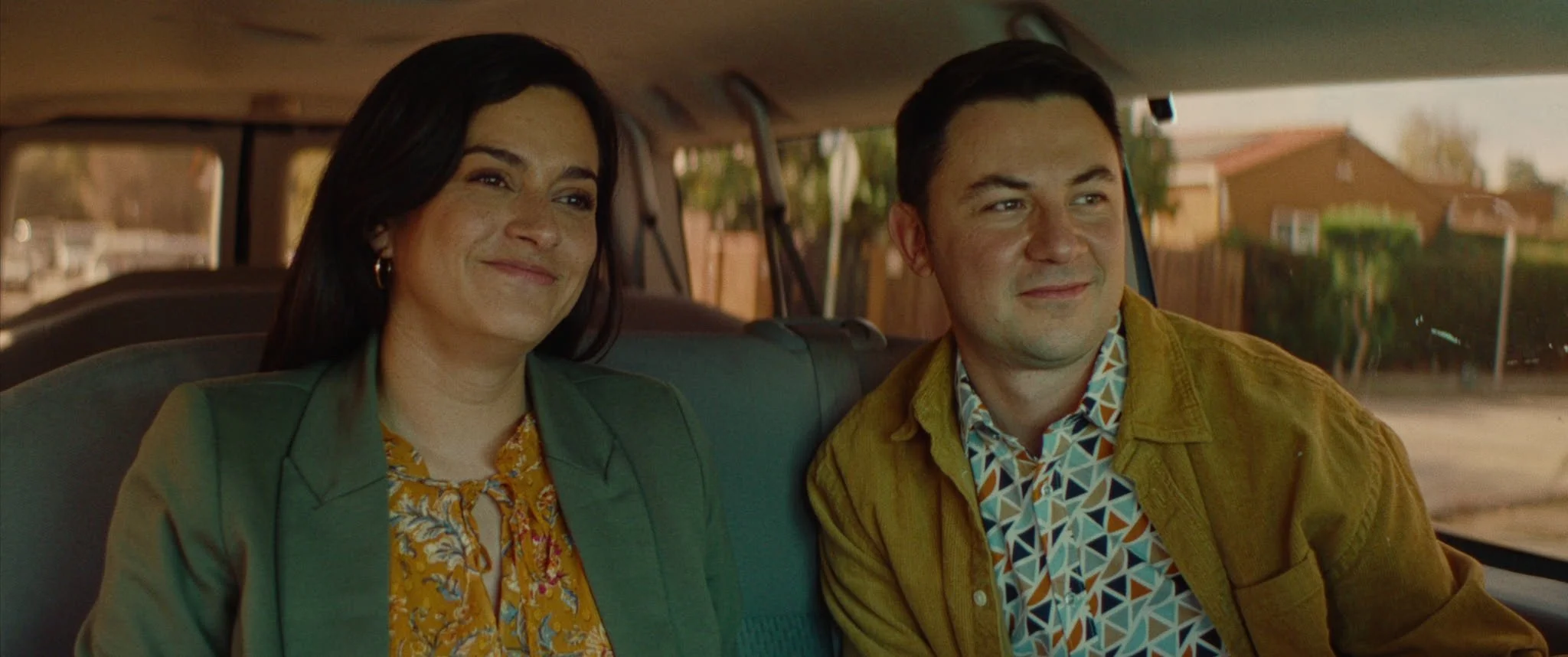 A brother and sister smiling side by side in the back seat of a family van.